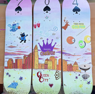 Hand-painted pastel skateboard decks featuring playing card symbols, whimsical illustrations, and Charlotte city skyline details on a soft lavender and gradient background.