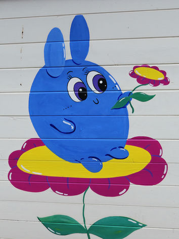 A colorful mural painted on a front gate featuring a giant blue bunny-like monster sitting atop a pink flower. The gate is covered in bright, playful colors and whimsical details, transforming an ordinary white gate into a cheerful outdoor artwork. The mural reflects a childlike, imaginative style with bold shapes and vibrant hues.