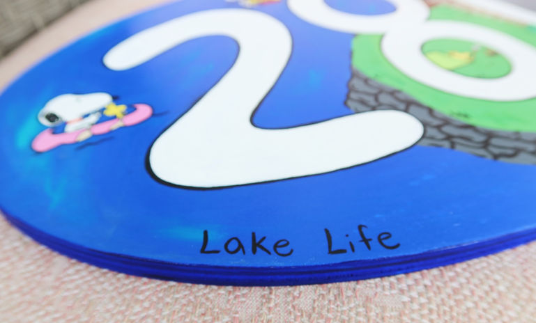 A colorful acrylic painting of a peaceful lakeside scene featuring a wooden dock, calm blue water, and lush green trees. An oval wooden sign displaying the number "280" sits in the foreground, representing a family lake house. Tiny Snoopy characters are hidden throughout the landscape, adding playful, nostalgic details that celebrate childhood memories, family traditions, and summers spent at the lake.