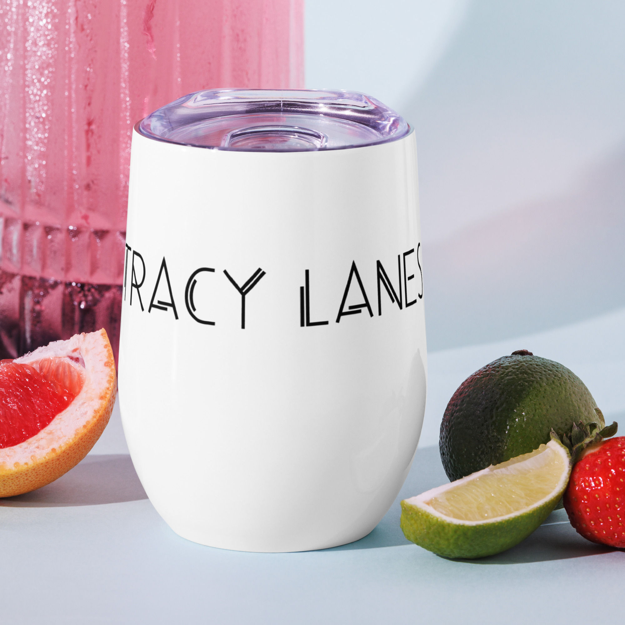 White tumbler with the text "TRACY LANES," next to fresh fruit and a pink beverage container in the background.