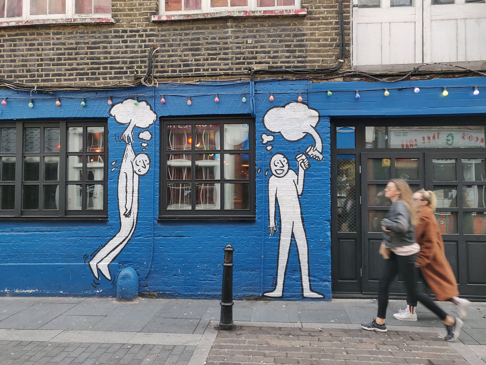 Two women walk past a blue brick wall with cartoon murals. One figure holds another by its head, both with thought bubbles. Colorful lights above.
