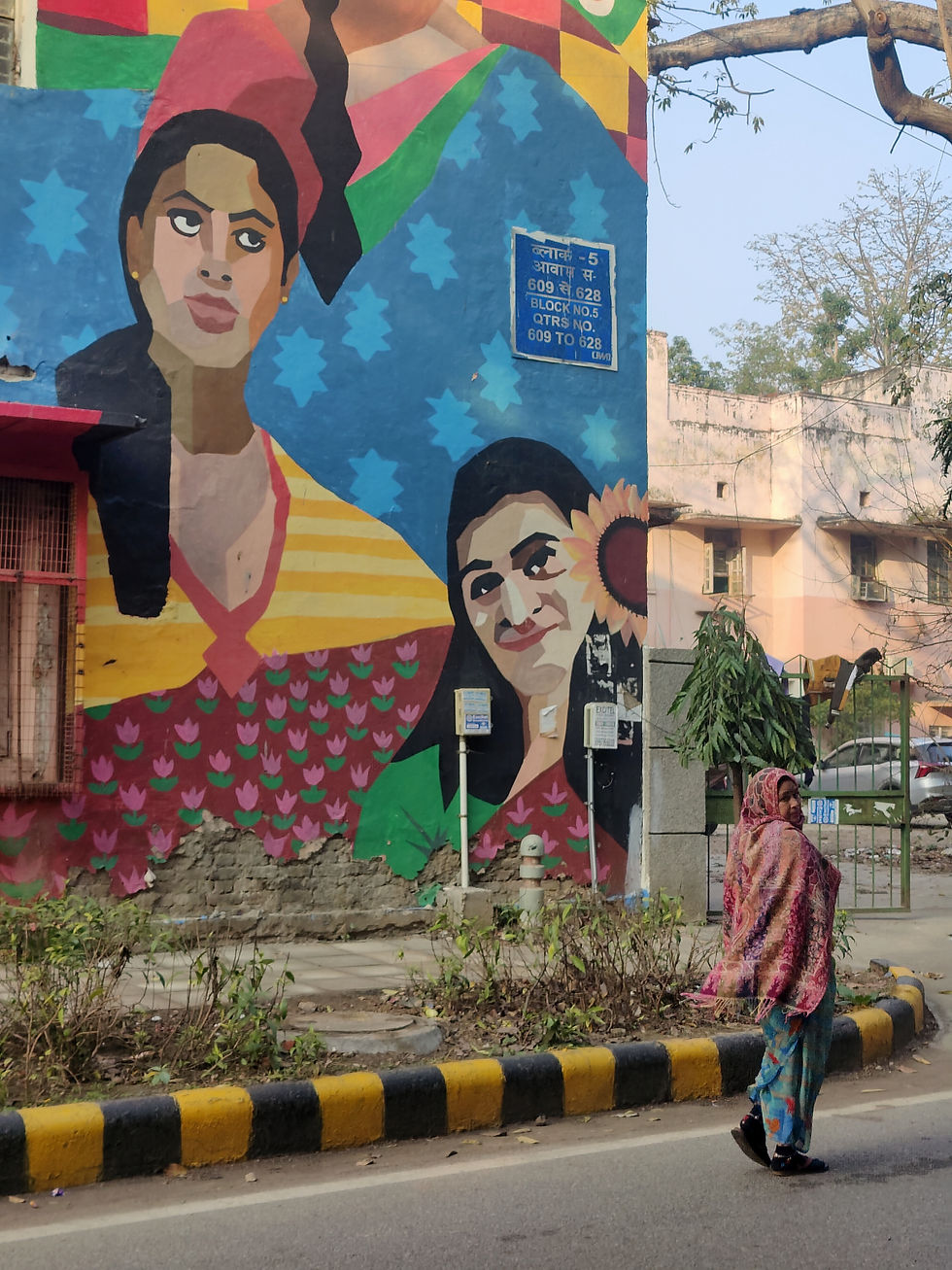 A woman in colorful attire walks past a vibrant mural of two women on a building wall. The mural features bold patterns and a blue sign.