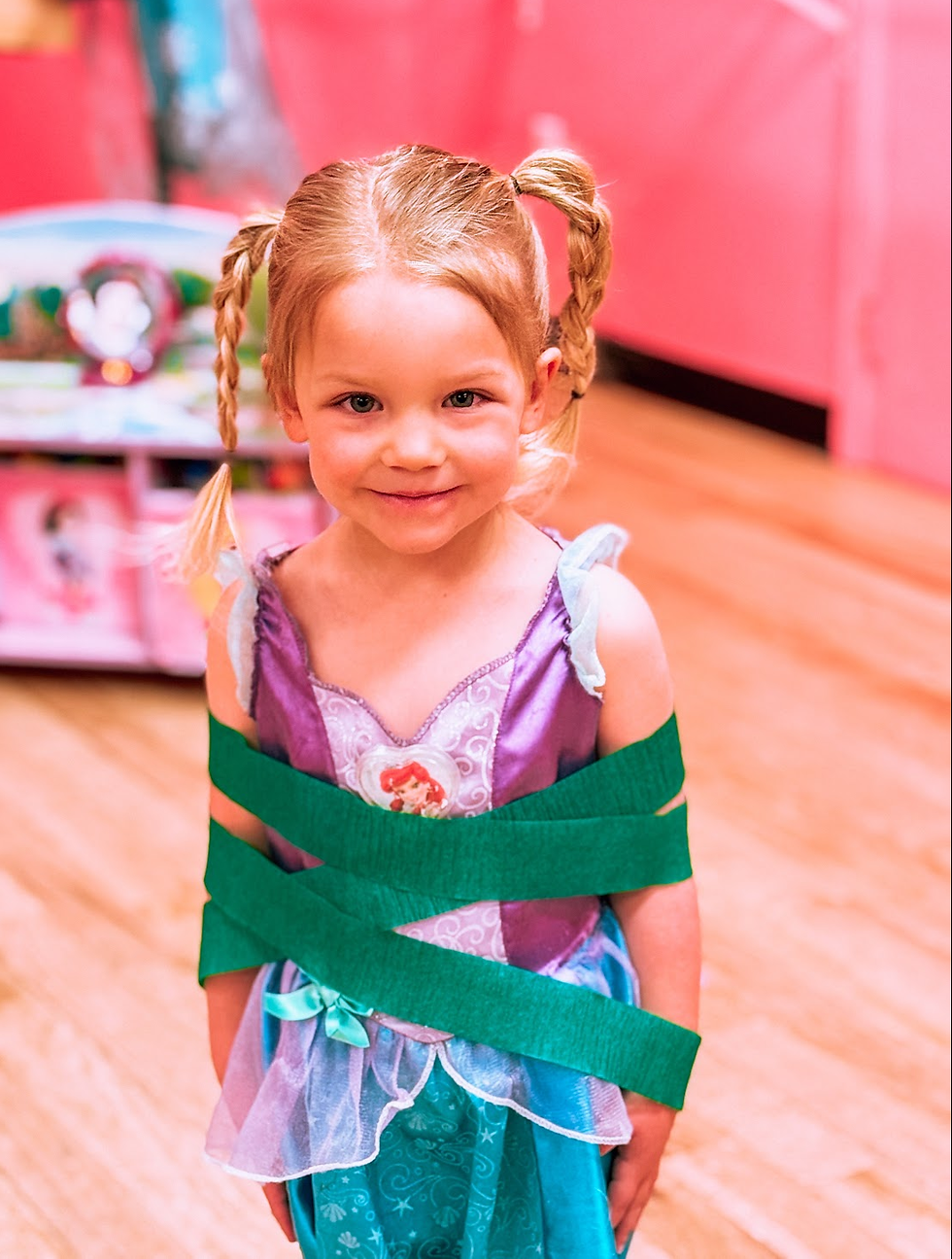 A young girl in a princess dress smiles as she is wrapped up in green streamers