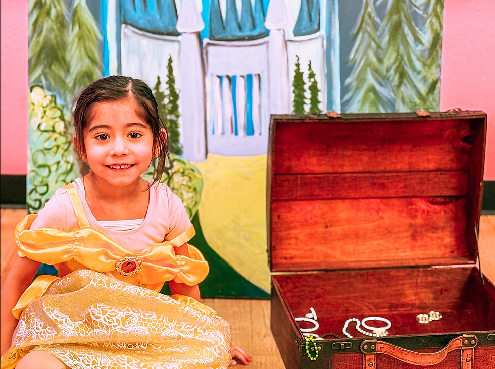 A girl in a Princess Dress smiles and poses in front of a princess castle and next to a treasure chest