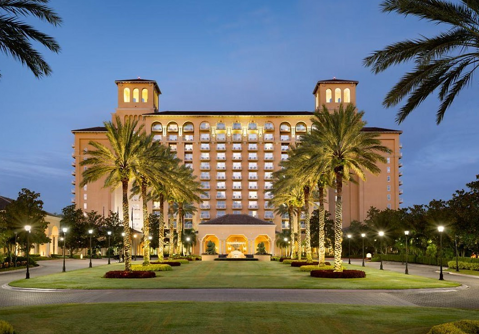 Elegant hotel with palm trees in a landscaped garden, lit warmly against a twilight sky. Arched windows and symmetrical design create grandeur.