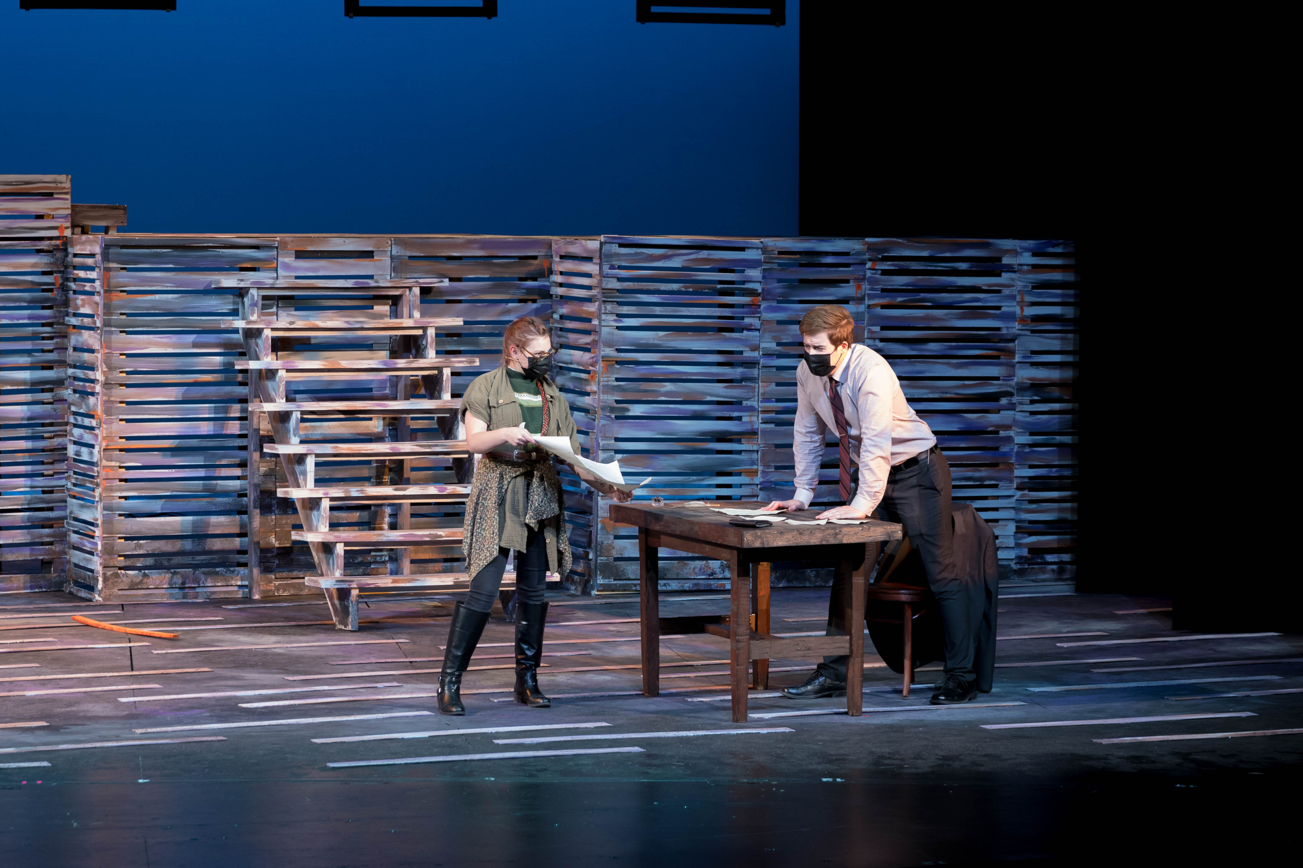 Two people on a stage under blue lighting, on opposite sides of a desk and going over papers.