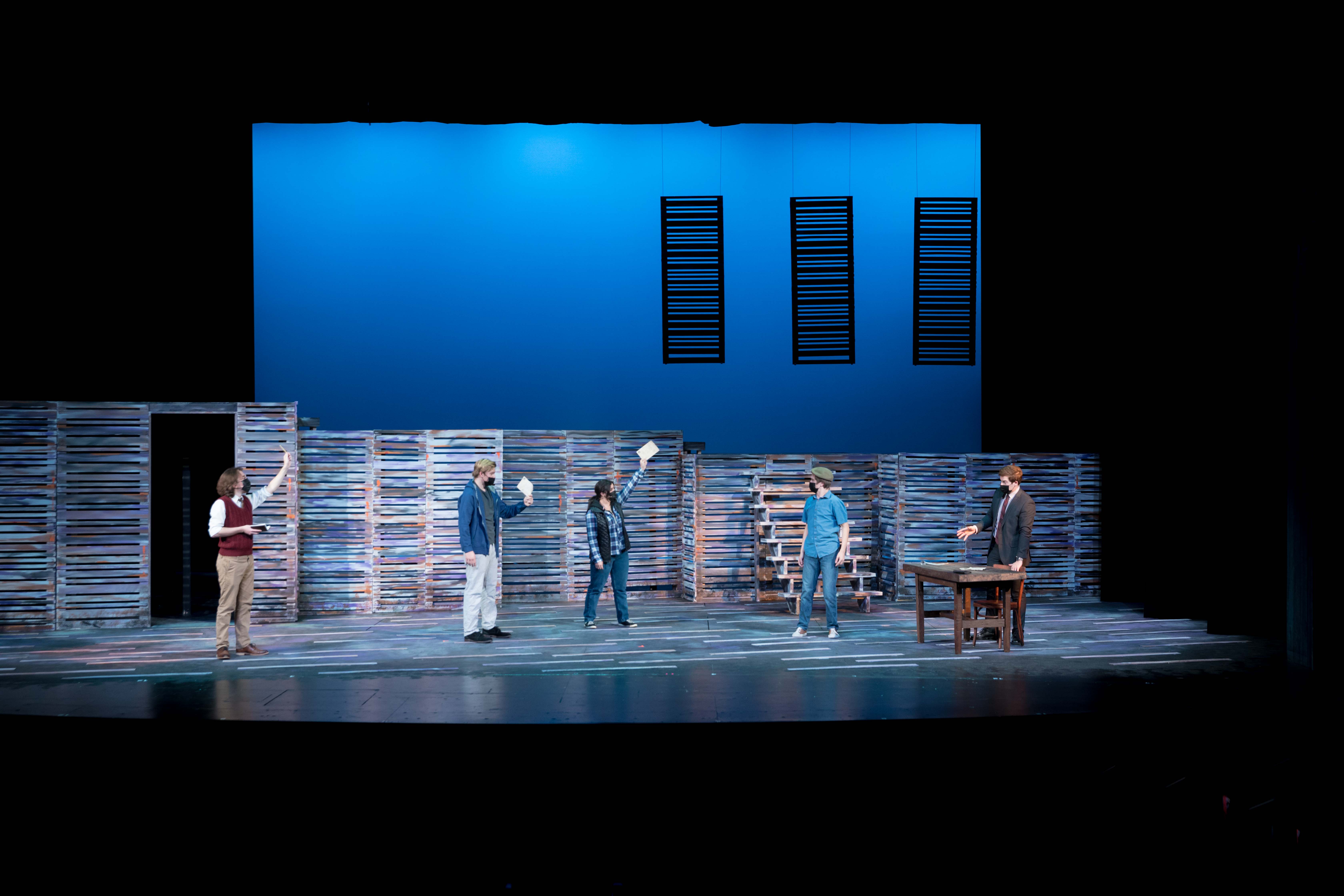 Five people on a stage under blue lighting, raising their arms and holding paper while one person stands behind a desk.