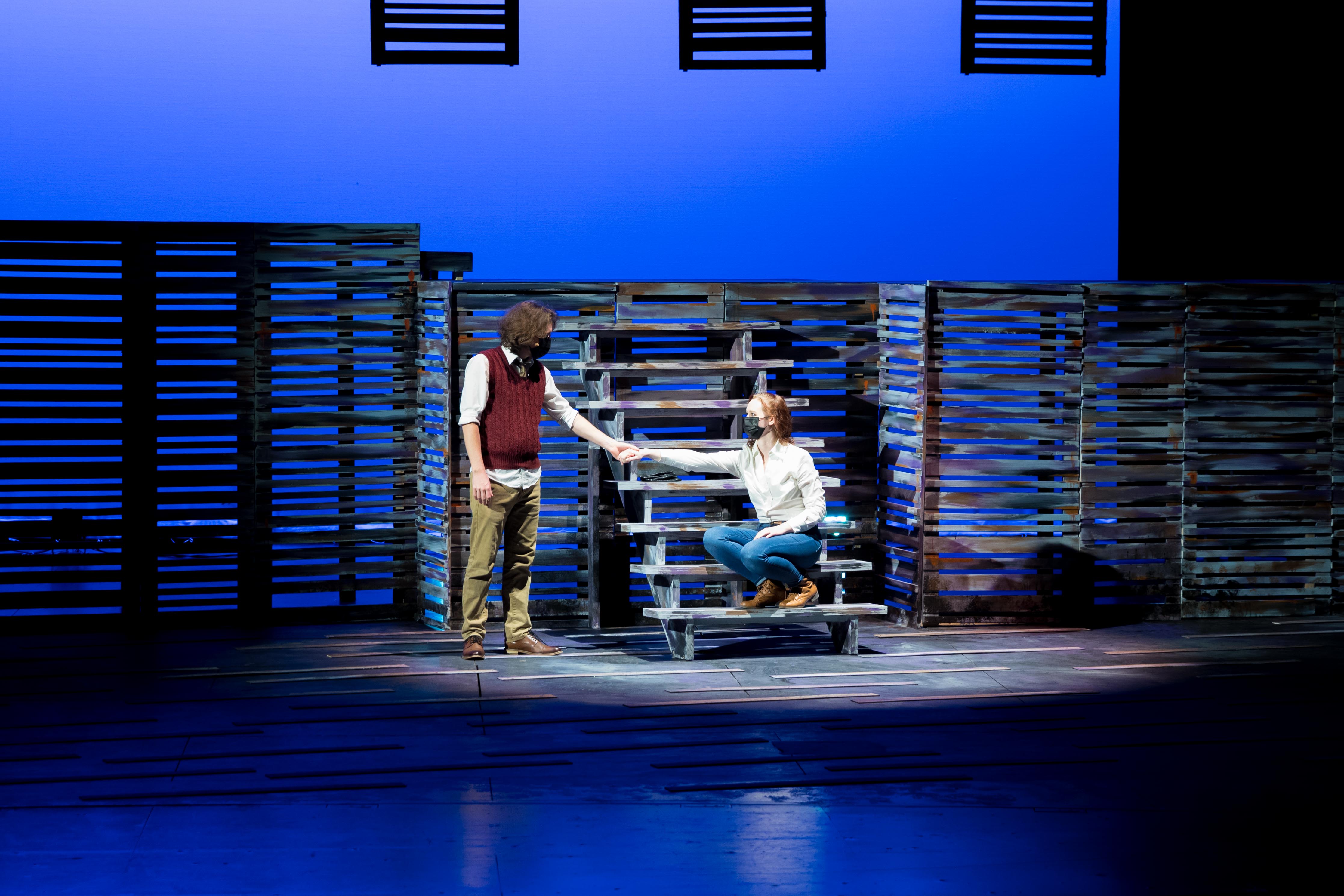 Two people on a stage under blue lights and a spotlight, one sitting on a staircase, both holding hands.