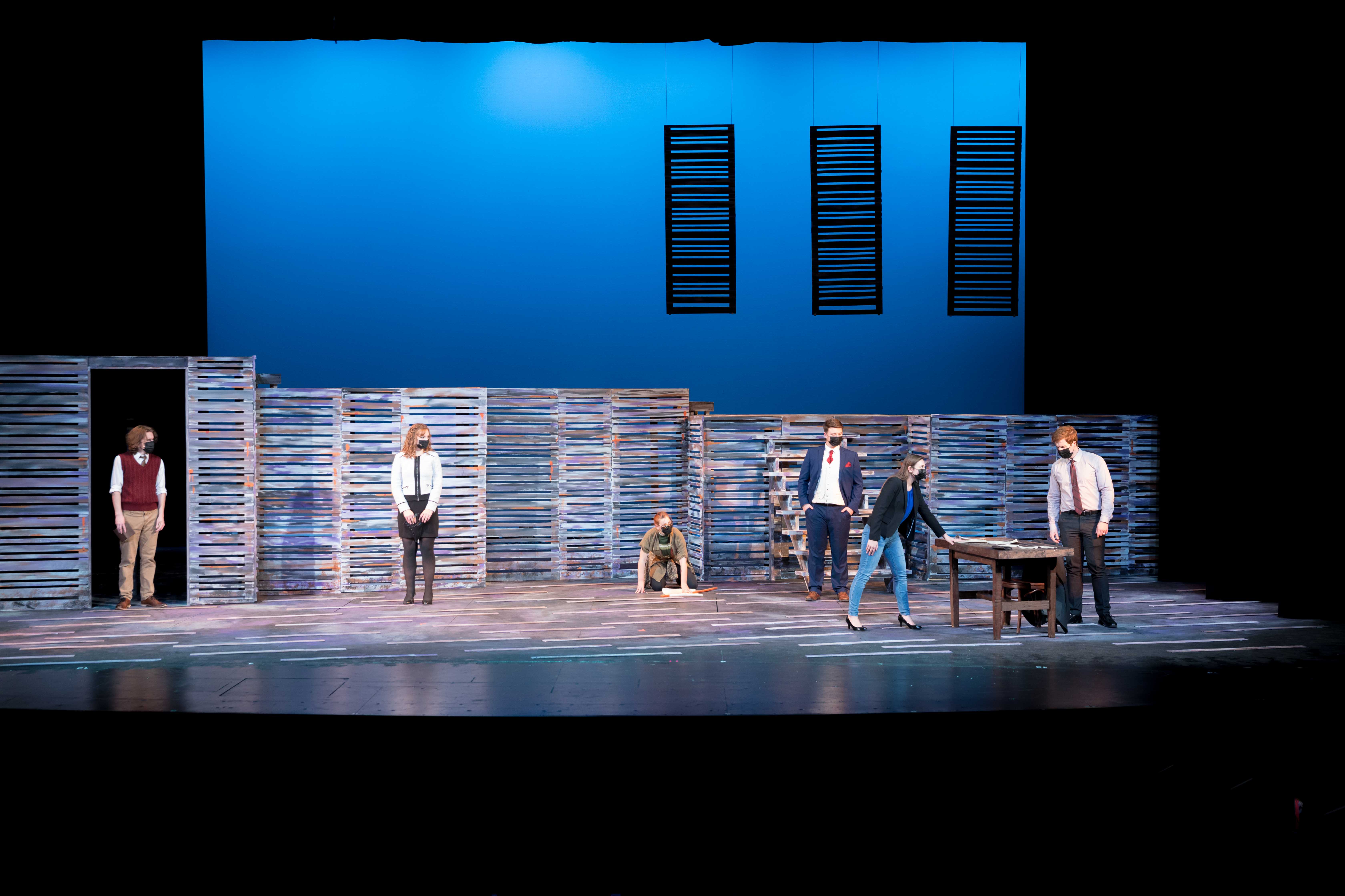 A group of people on a stage under blue lighting, one person behind a desk and one person crouched on the ground with papers.