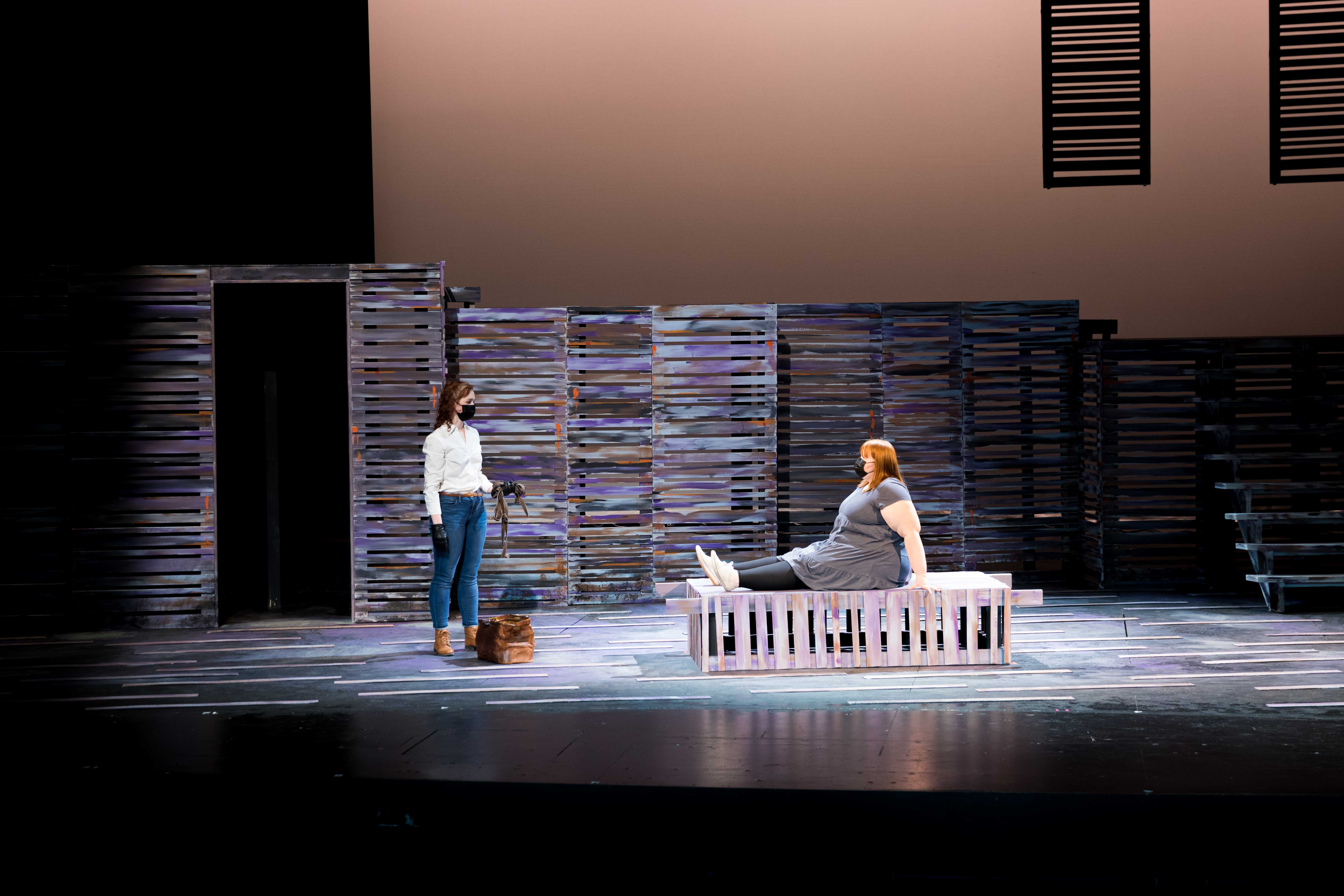 Two people on a stage under pink light, one standing with a bag, one laying on a bed.