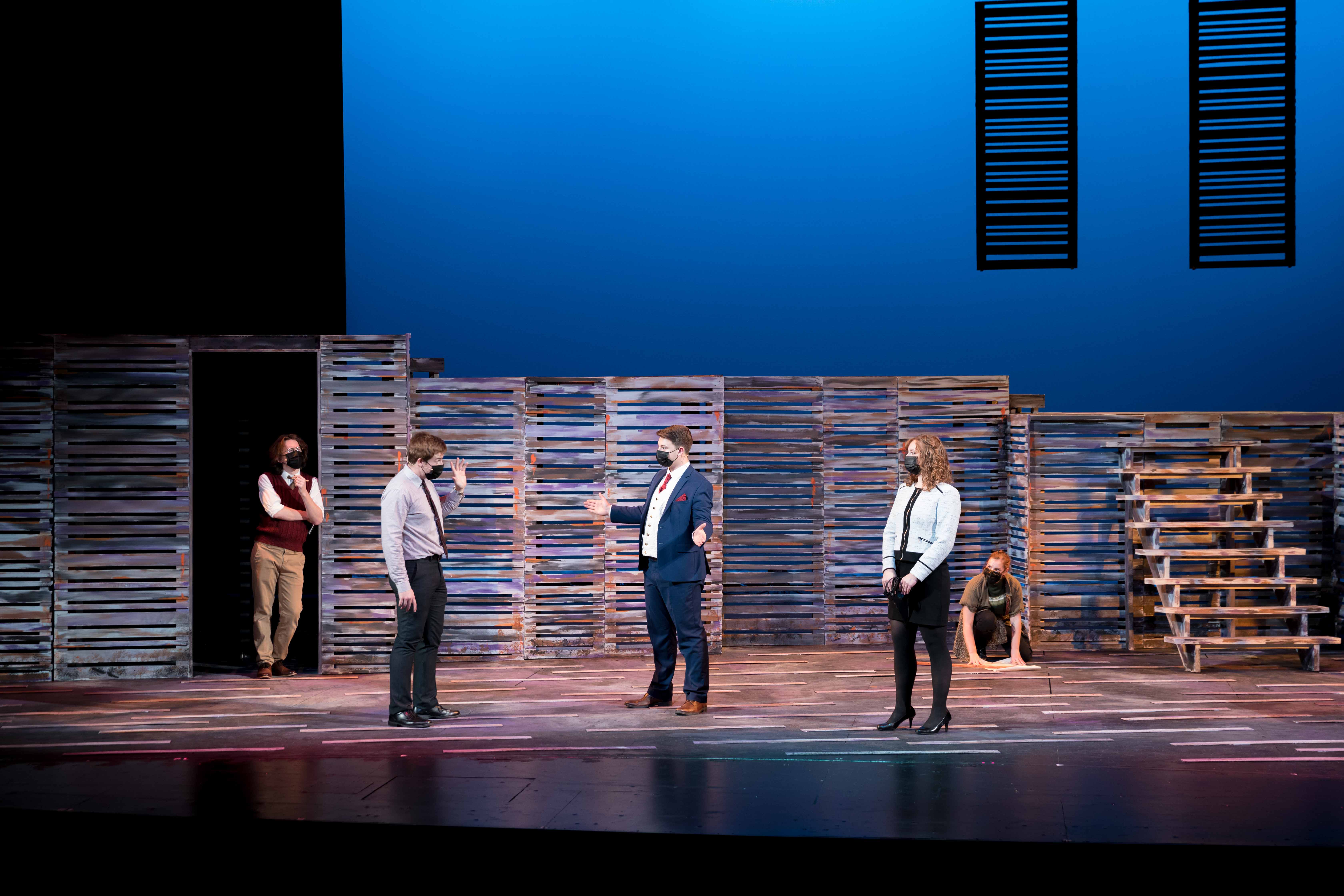 Five people on a stage under blue lighting, with one person crouched on the ground with papers.