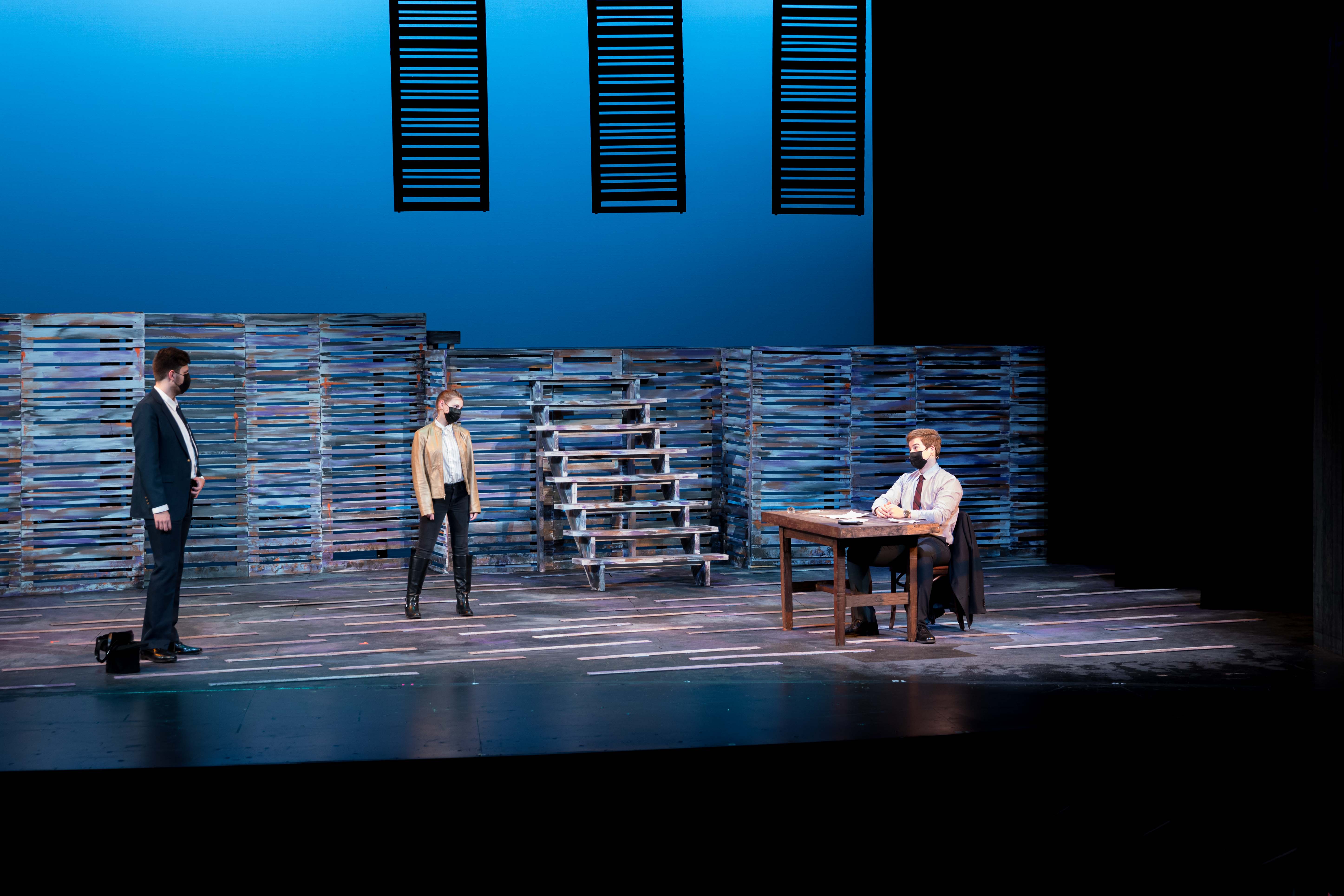 Three people on a stage under blue light, one behind a desk with papers.