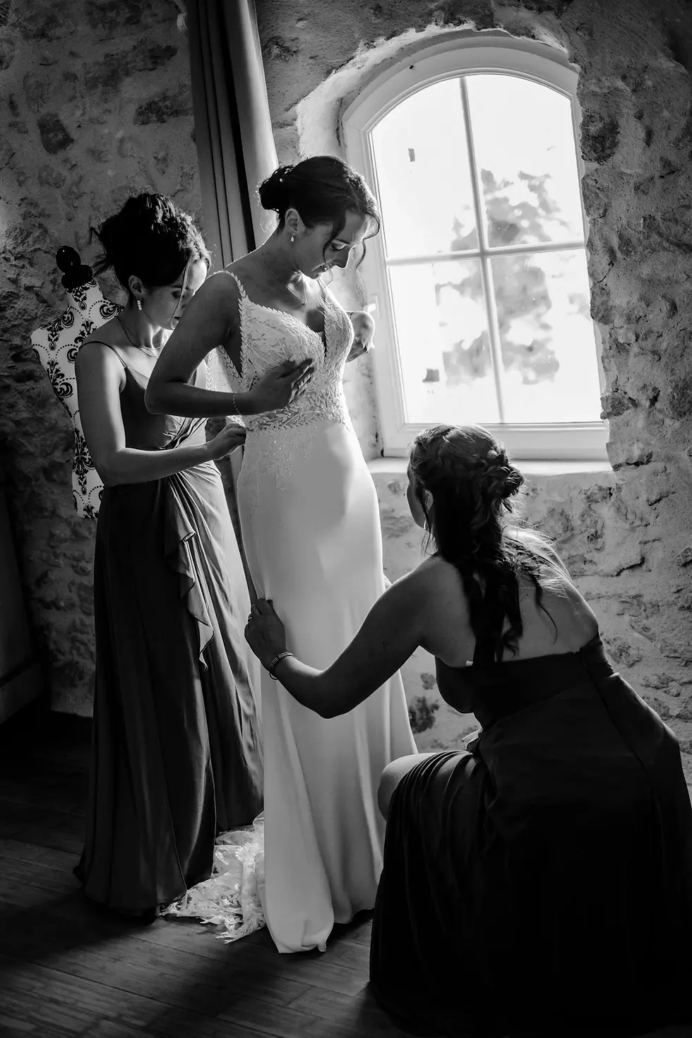 alt="Les demoiselles d'honneur aident la mariée à enfiler sa robe devant une fenêtre voûtée lors d'un mariage en Gironde"