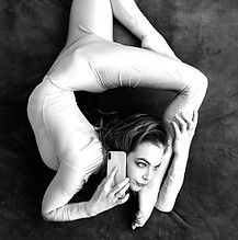 A female acrobat lies on a gray bedspread in a curved pose and uses a mobile. A woman cont