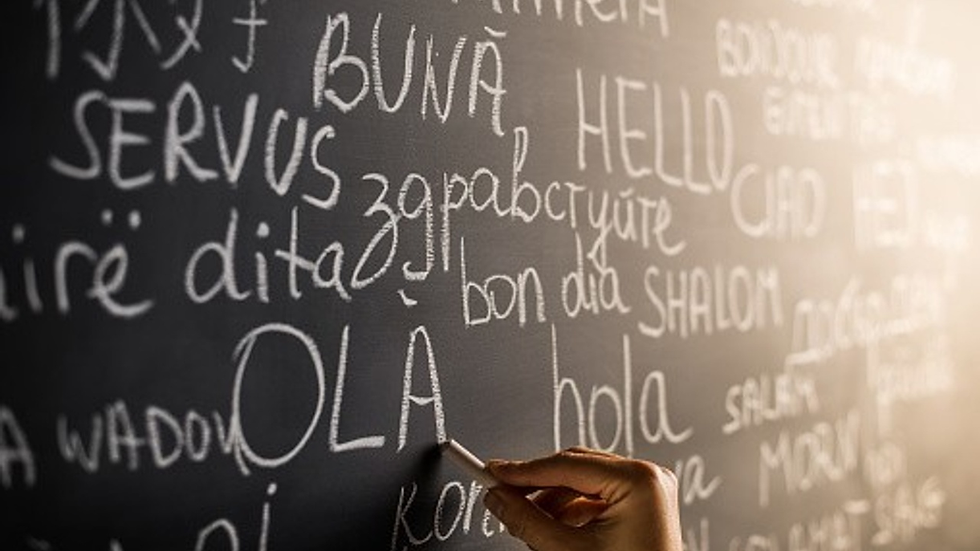 A close-up of a chalkboard covered with greetings written in multiple languages, including “hola,” “bonjour,” and “shalom,” as a hand writes the word “OLA” in chalk, symbolizing language learning and multilingual communication.