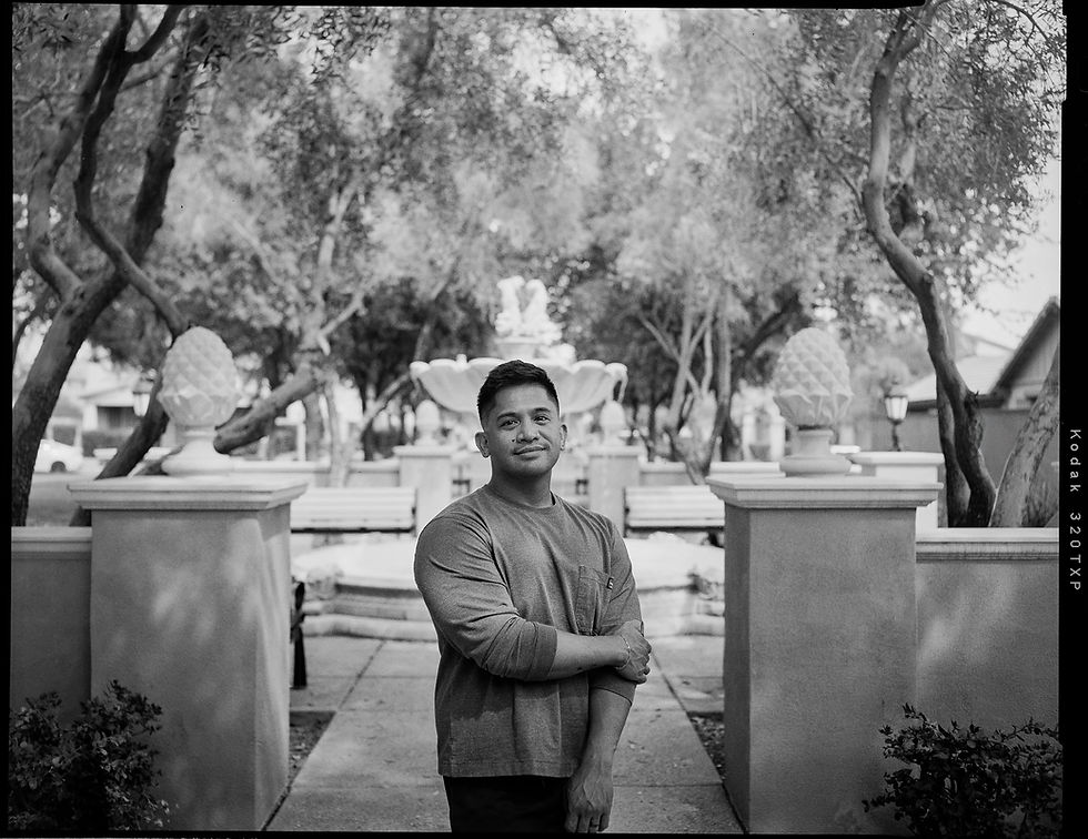 An authentic black and white portrait of a man standing by a fountain in Verrado, Buckeye, Arizona, arms crossed with a slight smile. Captured on 4x5 Kodak Tri-X 320 film using a vintage Busch Pressman press camera.