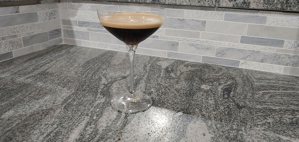 Dark espresso martini in a coupe glass on a gray granite kitchen counter, with a tiled backsplash behind it.
