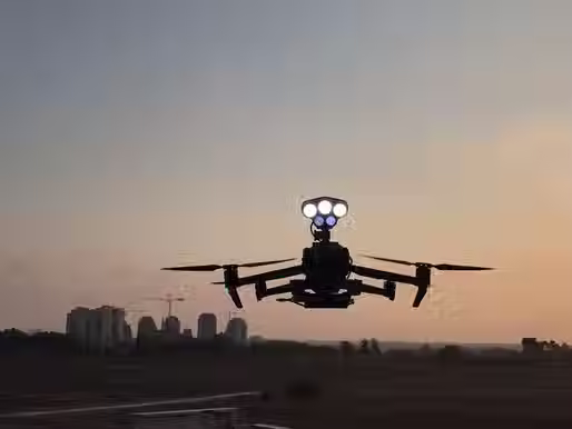 A DJI Mavic 2 drone with Drone Sky Hook searchlights seen hovering above a city with a sunset background