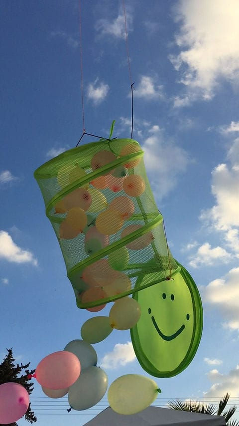 Green mesh basket with smiley face hangs in blue sky, filled with colorful balloons. Balloons cascade out, creating a playful scene.