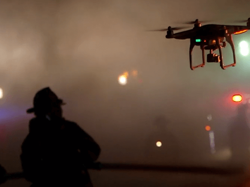 Firefighters in silhouette hold a hose amid smoke. A drone hovers nearby with flashing lights. Emergency vehicles are blurred in the background.