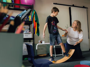 A child balances on a curved wooden balance board indoors while an adult kneels nearby, offering steady support in a therapy room with sensory equipment and shelves in the background.