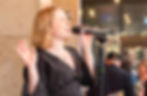 A woman sings passionately into a microphone, wearing a black dress, with string lights in the background setting an intimate mood.