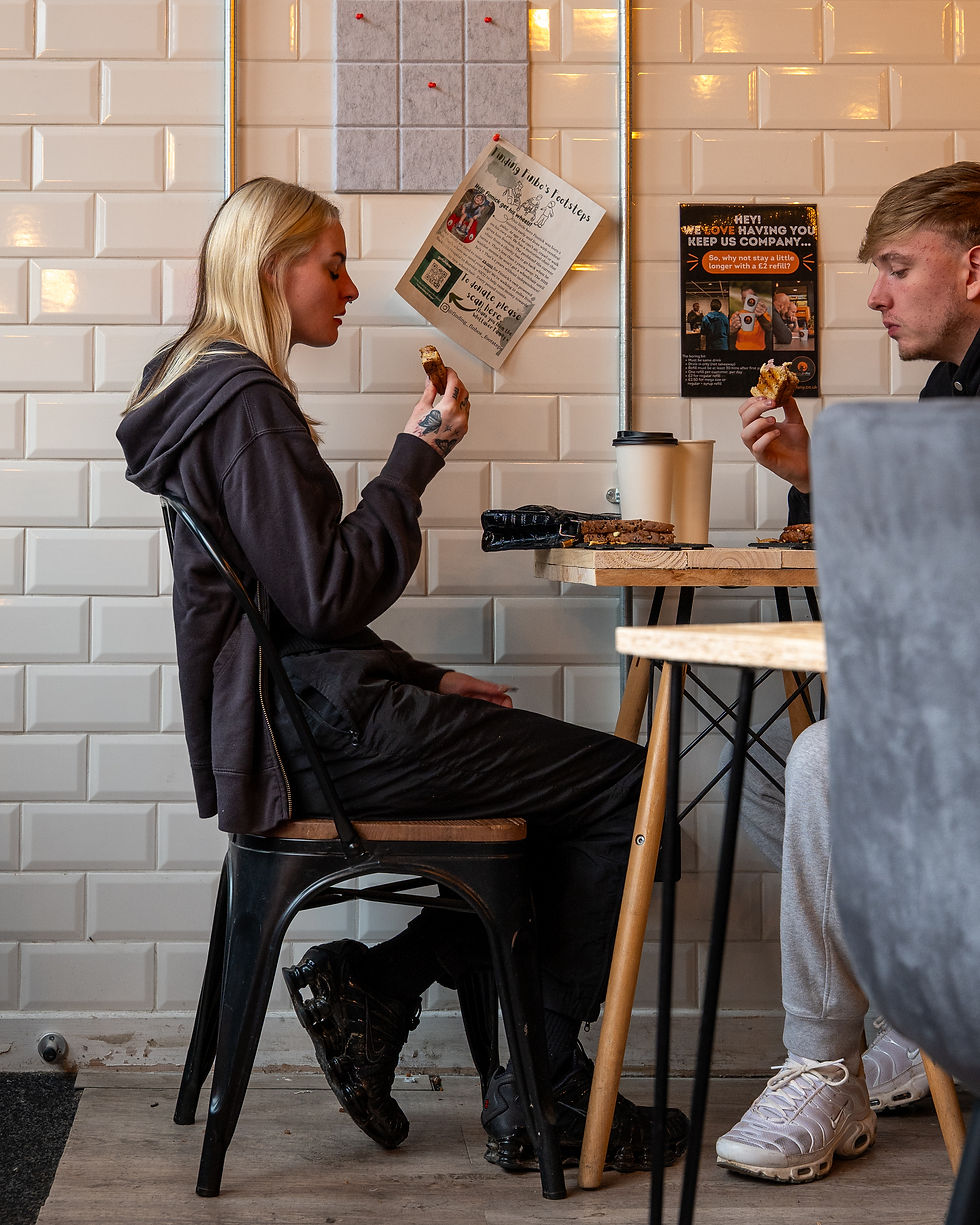 Two people sitting at a cafe table eating sandwiches. The background has white tiled walls with posters. A cup of coffee is on the table.