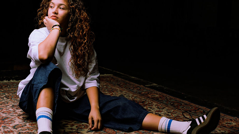 Young person with curly hair sits on a patterned rug, wearing a white shirt and jeans, looking thoughtful in a dimly lit room.