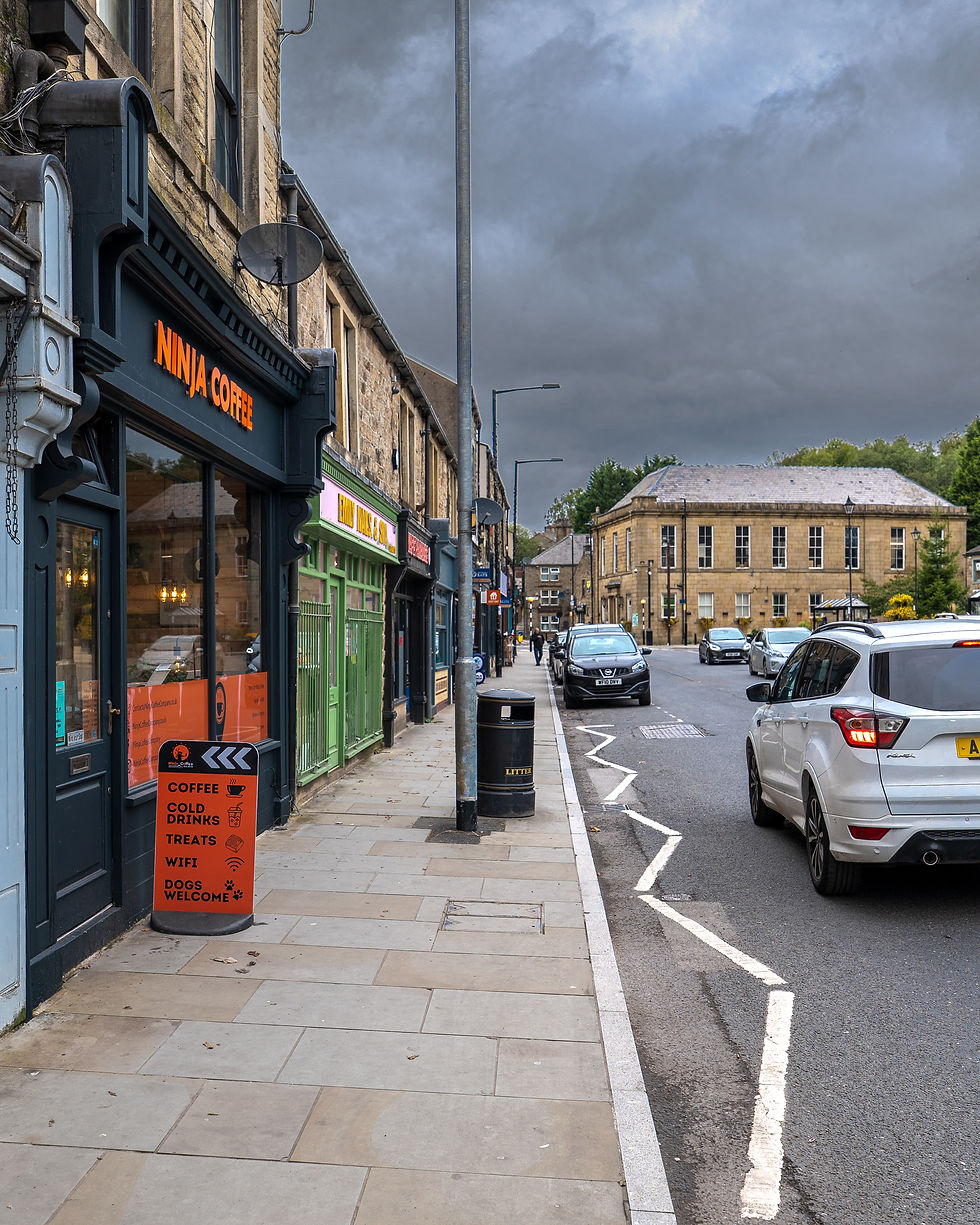 Street with shops, including "Ninja Coffee." Cars parked on road, cloudy sky, and bright orange sign offering coffee and treats.