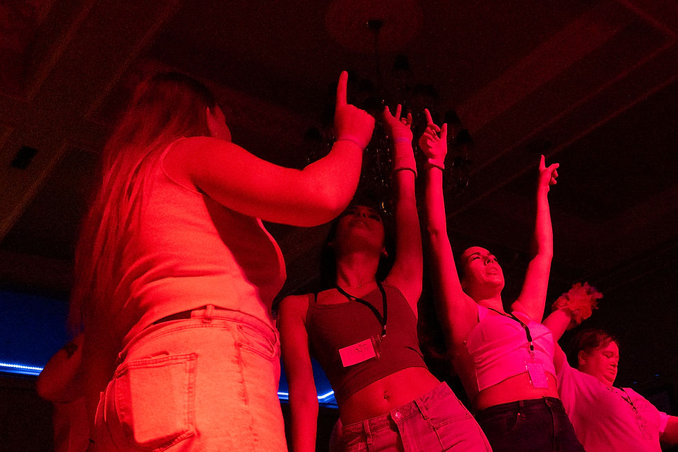 People dancing energetically under red lighting, hands raised. Name tags visible. Festive and lively atmosphere in a dimly lit room.