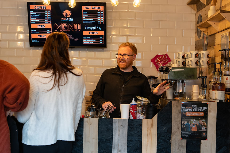 Barista with glasses smiles, talks to two women at counter. Coffee menu on wall, cups stacked, wooden and tiled decor. Warm atmosphere.