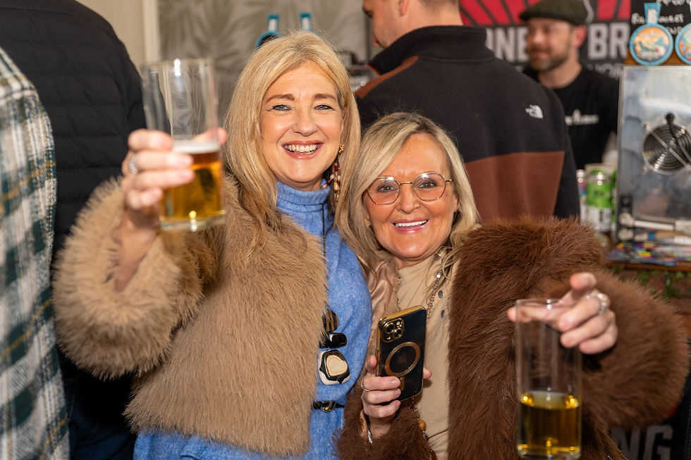 Two women in fur coats smiling and holding drinks at a lively bar. A man in the background wears a cap. Warm, cheerful atmosphere.