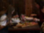 Two people seated at a wooden table, engaged in eating. Plates and a charcuterie board with bread, meat, and olives are visible. Rustic mood.