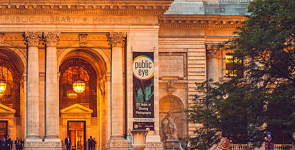New York Public Library at dusk with blurred pedestrians, lit entrance, skyscrapers behind, "Public Eye" banner, and orange cones on street.