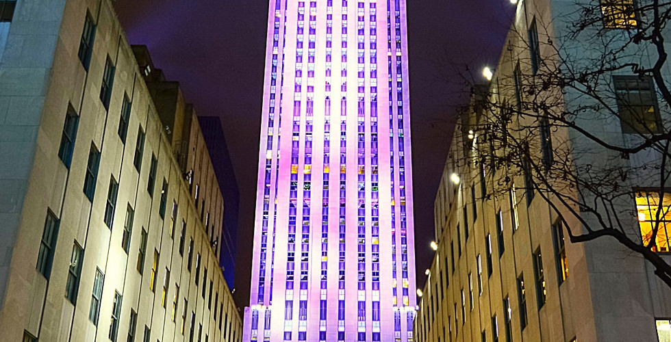 Tall building lit in purple towers over a crowd at night. Colorful Christmas tree below. Text: Rockefeller Center, New York.