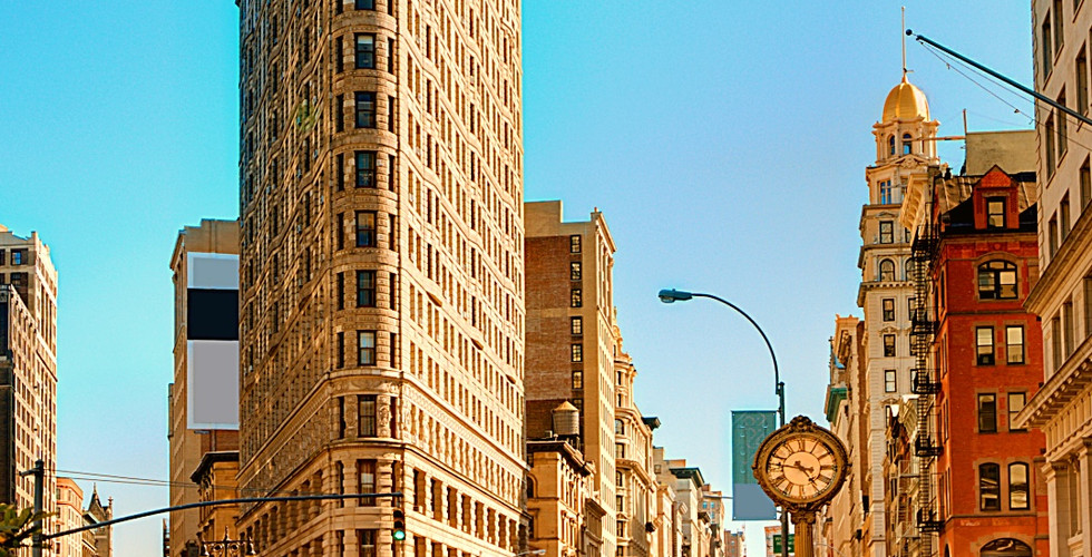 Flatiron Building under clear sky in NYC. Yellow cabs on busy street, tall historic architecture, vintage clock, lively urban scene.