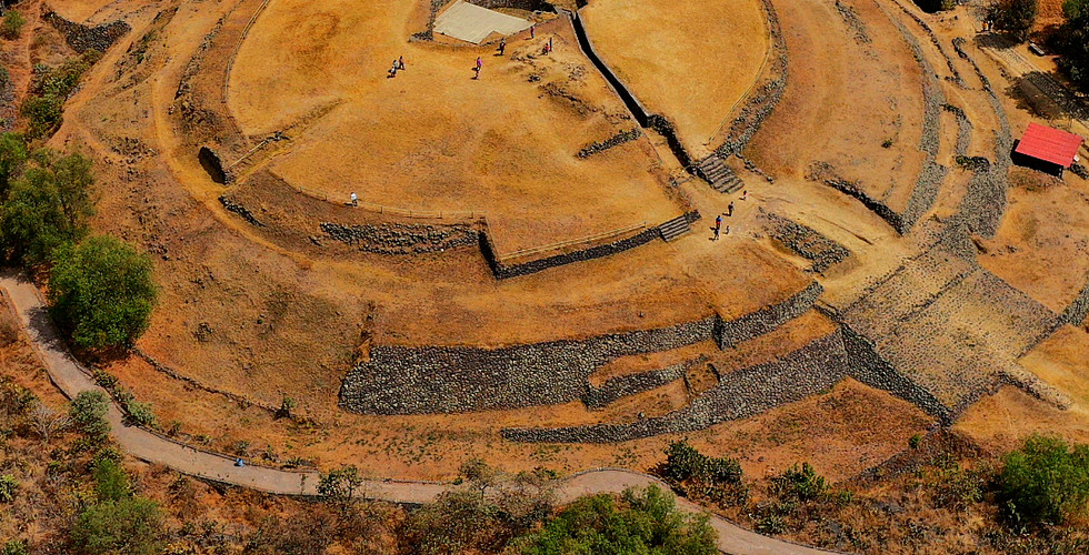 Aerial view of Cuicuilco, a circular ruins site in Mexico City, surrounded by trees and dry landscape. Visitors walking on site.