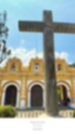 Yellow and white church facade with bells in Antigua, Guatemala. Foreground stone cross under blue sky. Text: Antigua Churches, coordinates.