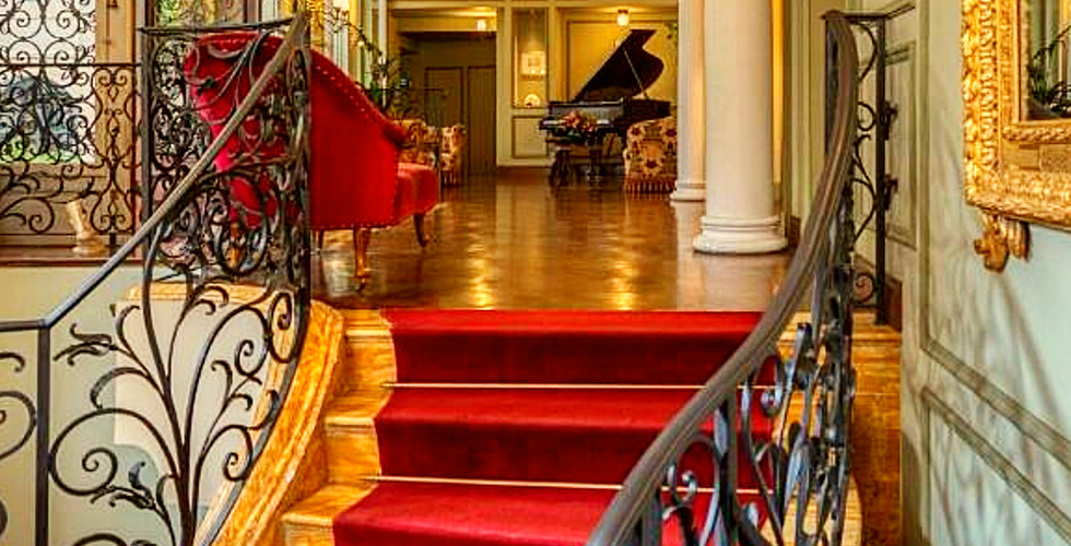 Elegant hotel interior with red-carpeted stairs, ornate iron railing. A grand piano is in the background. Gold-framed mirror on the wall.