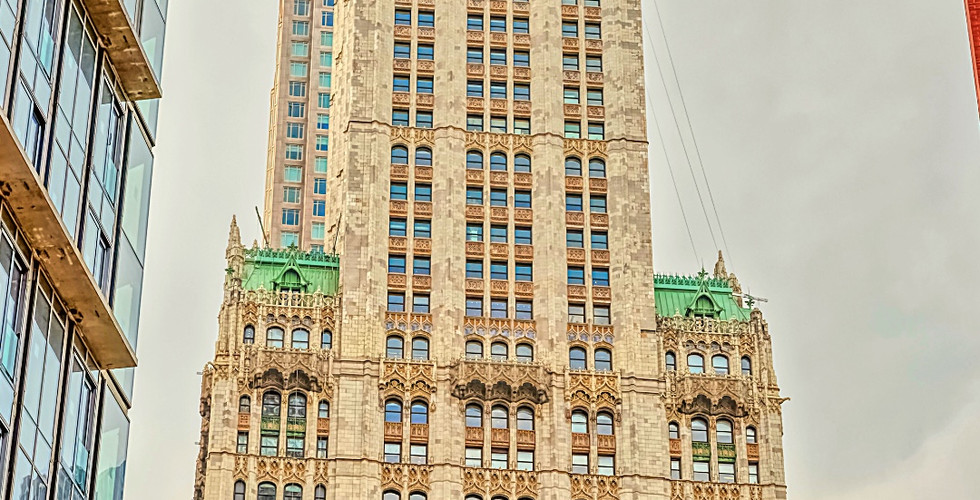 Tall skyscraper with ornate facade and green roof spire between modern buildings. Overcast sky. Text reads Woolworth Building, New York.
