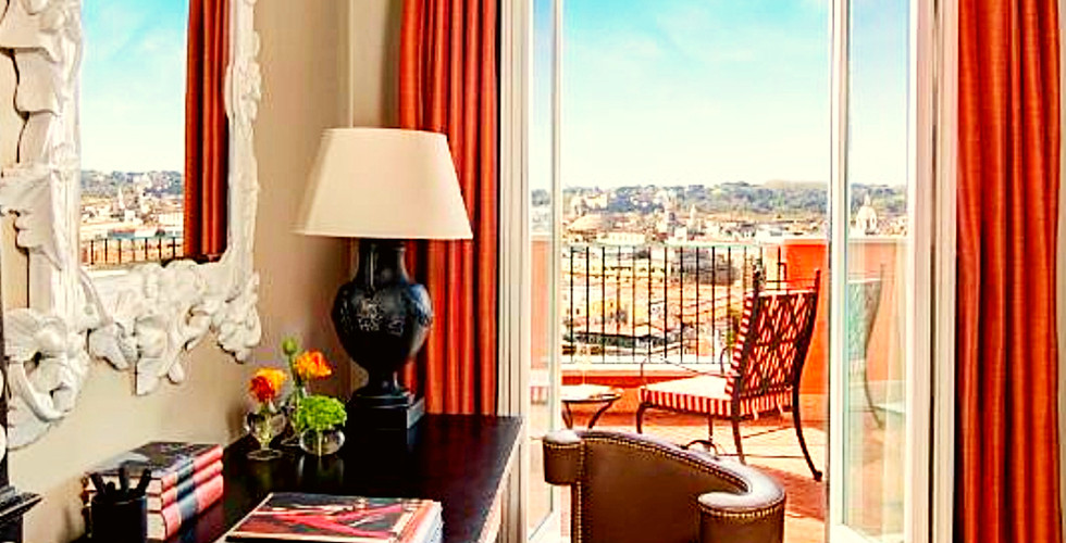 Sunny hotel suite with a desk, books, and lamp. Orange curtains frame open balcony doors. Cityscape view under blue sky. Calm ambiance.