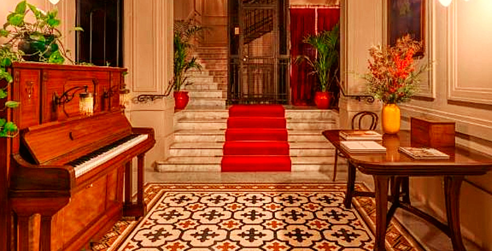 Elegant hotel foyer with chandelier, ornate tile floor, wooden piano, and red carpeted stairs. Warm lighting creates a cozy atmosphere.