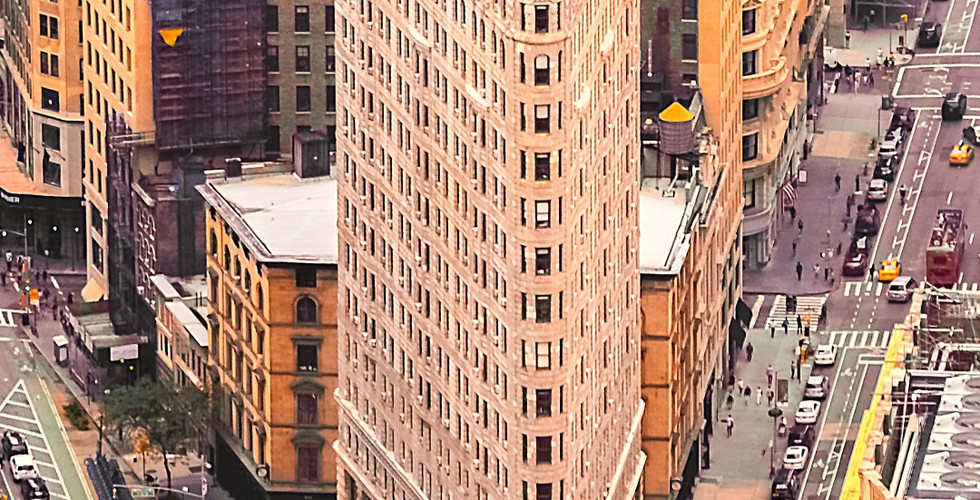Aerial view of the Flatiron Building in New York City, surrounded by yellow cabs and brown buildings. Text at bottom with location details.