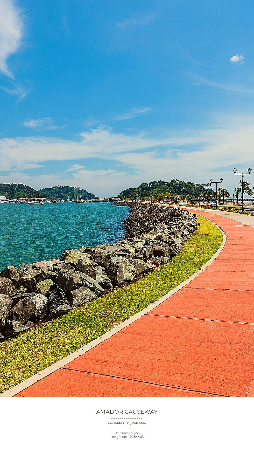Scenic view of Amador Causeway, Panama City. A red walkway curves beside blue water, rocky barrier, and palm-lined road under a clear sky.