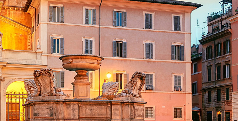 Square with a fountain in front of Basilica di Santa Maria in Trastevere at sunset. Warmly lit historic buildings and vibrant sky. Text below: Basilica of Santa Maria in Trastevere, Rome, Italy.