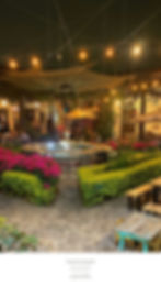 Courtyard with fountain, vibrant flowers, patio tables, and hanging lights at night. People dining in a lively, rustic atmosphere.