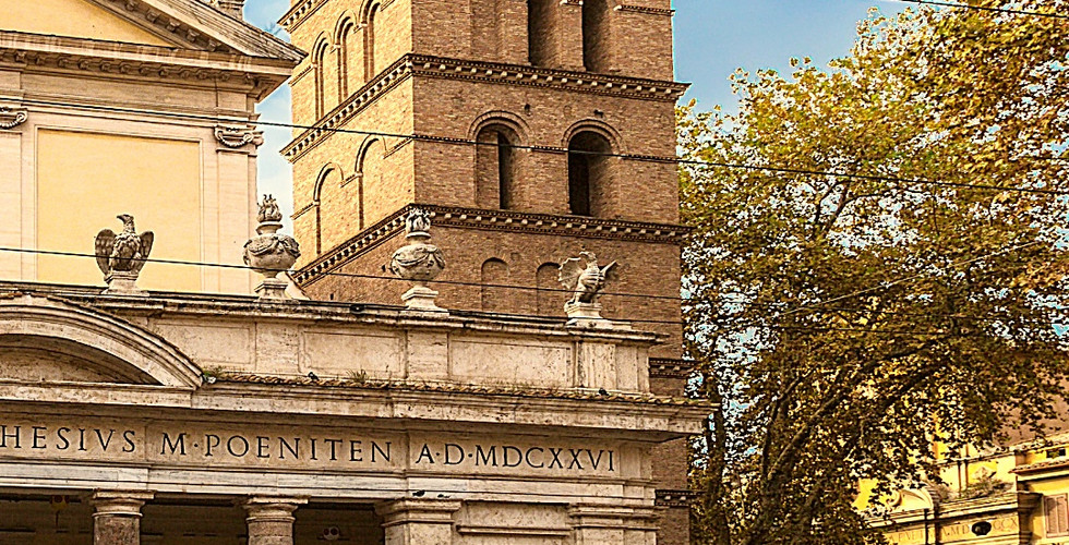 Basilica di San Crisogono in Rome, with a tall brick bell tower. Cars parked in front. Blue sky and trees in the background.