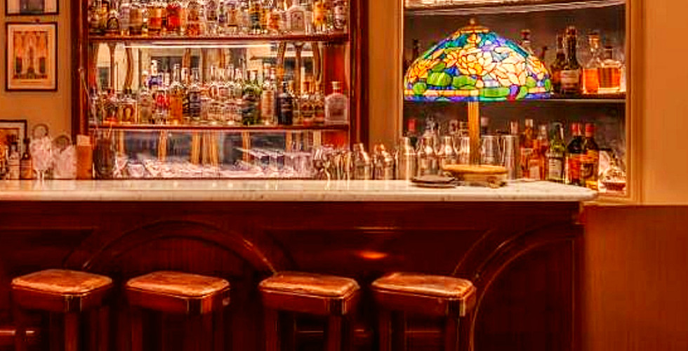 Elegant bar interior with stocked shelves, warm lighting, and a decorative lamp. Text reads "Hotel Locarno Bar, Rome, Italy." Cozy ambiance.