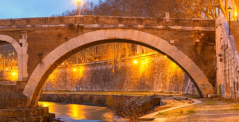 Bridge over a calm river at dusk in Rome, Italy, under a clear sky. Streetlights cast a warm glow on the stone path and surrounding trees.
