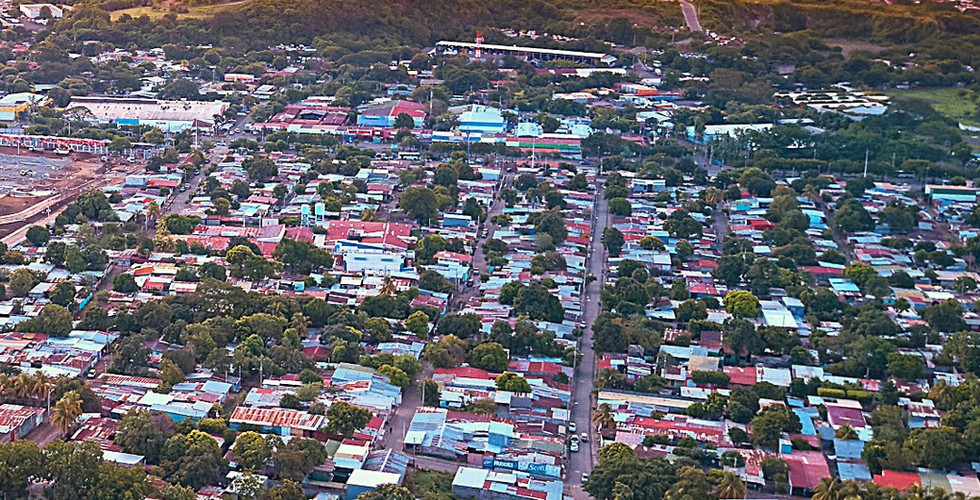 Aerial view of Managua at sunset with colourful rooftops and lush trees. Volcano in distance. Text: "Managua, Nicaragua. Latitude: 12.13, Longitude: -86.23."